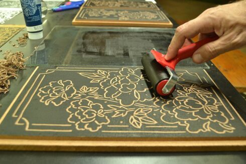 Xilogravura Peônias - Detalhes do Processo de Impressão, aplicando tinta sobre a matriz. Flores Peônia com moldura ao fundo. Materiais: Xilogravura / Gravura em madeira.
